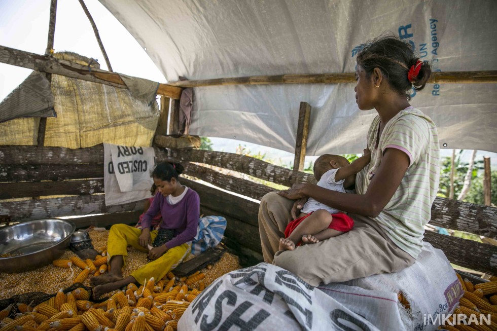 A mother sits on the corn bags breastfeeding her child while her other daughter grinds the corns to get the seeds out.